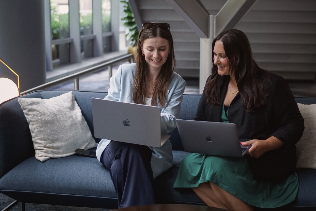 Two women sitting on a sofa in a corporate setting, each holding and looking at their laptops. They are in mid-discussion.