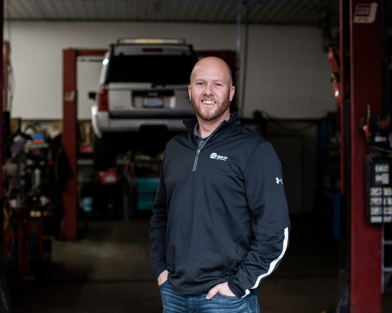 A man stands smiling inside an auto shop. There is a car being worked on in the background.