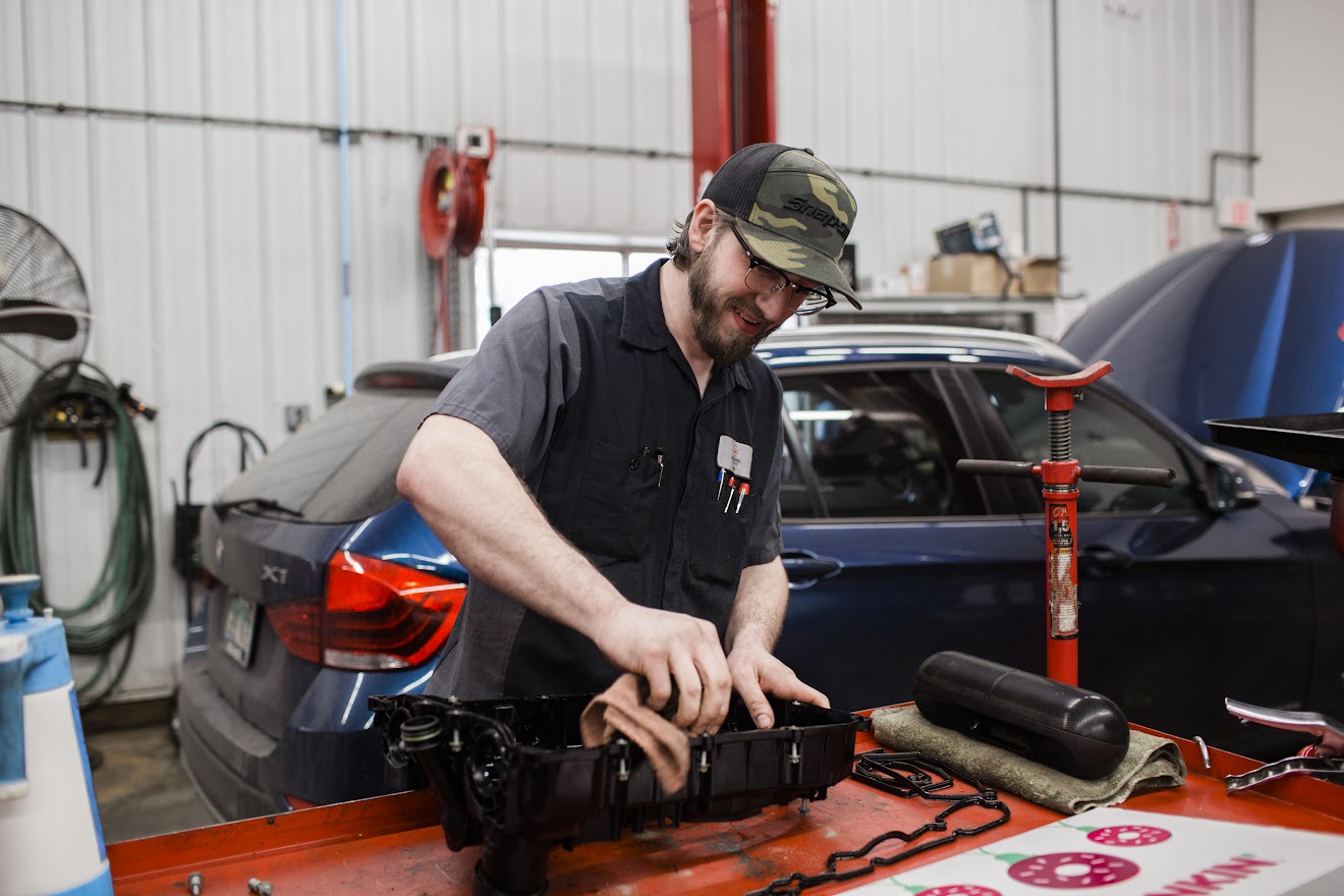 A man is working on a car, wiping a car part with a rag. He is in a mechanic shop and is smiling while looking at what he is doing.